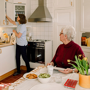 En äldre kvinna sitter vid sitt köksbord. En kvinna från hemtjänsten plockar i ett av köksskåpen.