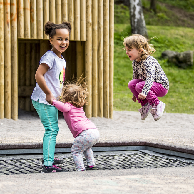 Bilden visar tre barn i olika åldrar som hoppar studsmatta på en lekplats.